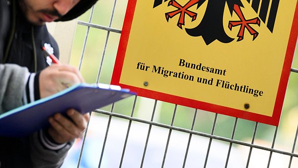 Ein Mann füllt ein Formular aus neben einem Schild vom Bundesamt für Migration und Flüchtlinge. Die Mitarbeiter der Behörde entscheiden über Asylanträge. (Archivbild) - © Julian Stratenschulte/dpa