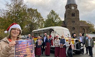Karin Heimke zeigt das Plakat für den Rehmer Weihnachtsmarkt. Im Hintergrund sind ihre Mitorganisatoren zu sehen, der Oldtimerbus und natürlich die Rehmer Kirche. - Nicole Sielermann