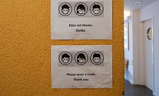 Hinweisschilder "Bitte mit Maske. Danke." hängen neben einem Eingang zu einer Arztpraxis. - picture alliance/dpa