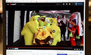 Sequenz mittendrin: Ein Filmausschnitt, der die Arbeit der Helfer unter erschwerten Bedingungen in Schutzanzügen zeigt. - Screenshot: Simone Flörke