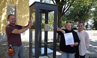 Michael Wieczorek (v. l.) von der Firma Art Medial bringt das Logo auf dem neuen Bücherschrank an. Regina Schlüter-Ruff und Melek Arslan von der Stadt Spenge schauen zu. - Mareike Patock