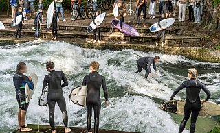 Surfer stehen am Eisbach in München. Zuschauer beobachten gespannt das Geschehen. - IMAGO/Wolfgang Maria Weber