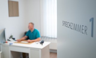 Ein Hausarzt sitzt in einem Sprechzimmer seiner Hausarztpraxis an einem Schreibtisch und arbeitet am Computer. - picture alliance/dpa