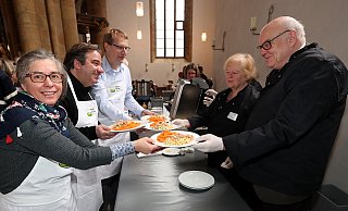 Ulrike Mann, Vincenzo Copertino, Joachim Hood (v.l.) tragen als ehrenamtliche Kellner weiße Schürzen mit Vesperkirchen-Logo und Namensschild. Wolfgang Ruwe und Anita Albers (v.r.) füllen an der Essensausgabe auf. - Wolfgang Rudolf