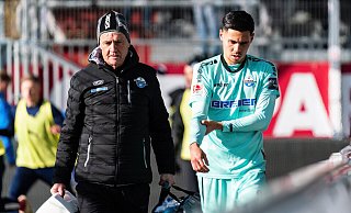 Raphael Obermair hält sich den lädierten rechten Arm und wird von Mannschaftsarzt Hans-Walter Hemmen in die Kabine des Holstein-Stadions begleitet. - Imago Images
