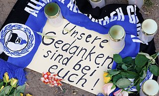 Kerzen, ein Fanschal von Arminia Bielefeld und ein Zettel mit der Aufschrift "Unsere Gedanken sind bei Euch!" sind am Tatort vor dem Club "Cutie" in der Innenstadt zu sehen. - Friso Gentsch/dpa