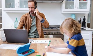 Ein Vater arbeitet zu Hause mit Smartphone und Laptop, während sein Sohn Schularbeiten macht. - picture alliance / Zoonar