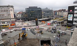 Mit Baggern wird jetzt die Hauptfläche vor der Niedernstraße aufgegraben (vorn). Rechts sind die neuen Haltestellendächer zu sehen – erstmals schon mit Grün obendrauf. Links am Pavillon gehen Passanten übers neue Pflaster. - Peter Unger