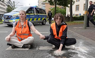 Mit einem besonders hartnäckigen Gemisch aus Klebstoff und Sand hat ein Klimakleber seine Hand auf dem Asphalt der Kreuzstraße befestigt. Ein Lösen mit Speiseöl ist unmöglich. Die andere Hand ist mit der seiner Mitstreiterin verklebt. - Paul Brinkmann