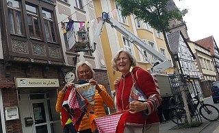 Eine von 70 Wimpelketten: Stadtwerke-Mitarbeiter Hannes Winter bringt vom Hubwagen aus die bunten Wimpel von Connie Tiemann (l.) und Monika Niemann über der Hauptstraße an. Rund 2.000 Exemplare hatten sie zusammen mit der Nähgruppe der "Zweiten Heimat" genäht. - Dieter Scholz
