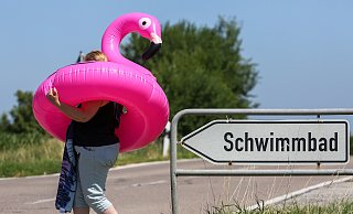Eine Frau läuft mit einem aufblasbaren Flamingo-Schwimmring und einem Badetuch an einem Wegweiser vorbei, auf dem "Schwimmbad" geschrieben steht. - Thomas Warnack/dpa