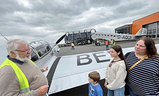 Hubert Schmidt vom Quax-Verein erzählt Levi, Lia und Katrin Seven (v. l.) Details aus der Geschichte der BF 108 aus dem Jahr 1937. Im Hintergrund sind die Ju 52 der Lufthansa und der Quax-Hangar zu sehen. - Jens Reddeker