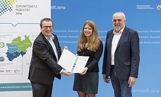 Verkehrsminister Oliver Krischer (l.) und Theo Jansen, Geschäftsstellenleiter vom Zukunftsnetz Mobilität NRW, übergeben Karolin Bludau nach erfolgreichem Abschluss in Düsseldorf eine Urkunde. - MUNV NRWRalph Sondermann