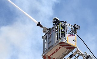 Ein Feuerwehrmann in voller Ausrüstung steht im Korb der Drehleiter, von wo er die Flammen auf dem Dach des Golden Tulip Hotel bekämpft. Die enorme Hitze von vorne, aber auch von oben und in der Kleidung lassen den Schweiß nur so fließen. - Barbara Franke