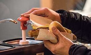 Ein Mann pumpt Senf auf eine Bratwurst im Brötchen. - dpa