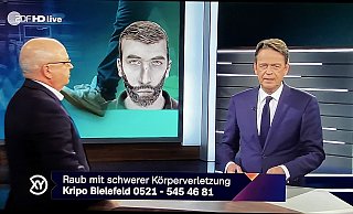 Kriminalhauptkommissar Thomas Grothaus gibt Rudi Cerne live im TV Auskunft zu dem Raubüberfall. Hinten ist das Phantomfoto des Täter s zu sehen. - Screenshot: ZDF / Aktenzeichen XY