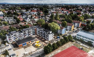 Links im Bild sind L-förmig die neuen Häuser zu sehen. Davor, orangefarben, der Treffpunkt mit seinen Beratungsstellen. Das aktuell schwarze Dach wird noch begrünt. Darauf folgen die Straße entlang drei Bestandshäuser der Genossenschaft sowie die Kita. Träger ist bei letzterer die AWO. - Gerald Dunkel