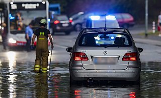 Duisburg: Ein Auto steckt im Hochwasser in einer Senke fest, während Feuerwehrleute die Gullydeckel öffnen, damit das Wasser ablaufen kann. - Christoph Reichwein
