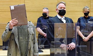 Ercan K, der sich hier am ersten Verhandlungstag eine Akte vor das Gesicht hält, steht wegen des Tatvorwurfs des zweifachen Mordes vor Gericht. - Barbara Franke