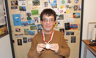 Benjamin Estermann präsentiert in seiner Wohnung die Goldmedaille. Er steht vor einer Wand, die mit Ansichtskarten beklebt ist, die aus der ganzen Welt stammen. Es sind Grüße von Tanzfreunden, mit denen Estermann in ständigem Kontakt steht. - Karsten Schulz