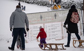 Ausflüger mit Schlitten gehen in Winterberg an einer geschlossenen Skipiste an einem Banner mit Corona-Verhaltensregeln vorbei.  - picture alliance/dpa