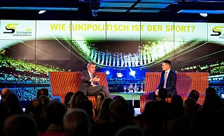 IOC-Präsident Thomas Bach (l) auf einer Bühne. Daneben sitzt Moderator Jens Zimmermann. - Tom Weller/dpa