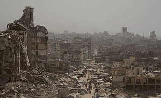 Menschen gehen eine Straße entlang, die von Gebäuden umgeben ist, die durch israelische Bombardierungen im Gazastreifen zerstört wurden. - Jehad Alshrafi/AP/dpa