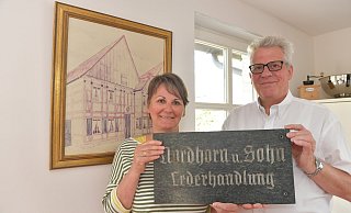 Andrea und Wilhelm Nordhorn halten das Firmenschild der 1910 von Wilhelms Großvater Franz gegründeten Lederhandlung. 1950 wurde der an der Königstraße ansässige Sitz (Bild im Hintergrund) der früheren Schäftemacherei nach Niehorst transloziert. Foto: Jens Dünhölter