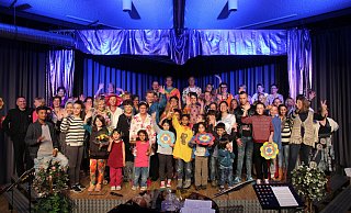 Das Bild zeigt den Chor der PaderVoices auf der Bühne gemeinsam mit ihren fröhlichsten Fans, den Flüchtlingskindern. Einige Kinder halten Prilblumen hoch, die zum Saalschmuck im Stil der Siebziger gehörten - xxxxxxxx