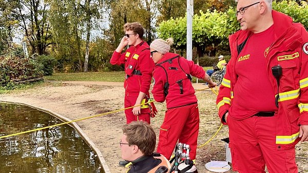An der Schnur sind die Signalgeberin Anna-Lena Mennecke und der stellvertretende Signalgeber Jonas Kahrmann. Sitzend am Ufer Adrian Koch, der Sicherungstaucher, der aktiv wird, sobald dem Einsatztaucher im Teich etwas zustößt. - © Robert Becker