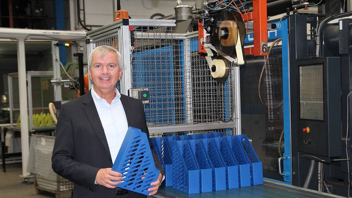 In der Produktionshalle: Geschäftsführer Ralf Niehaus steht vor einer sogenannten Spritzgussmaschine, in der die Büroartikel produziert werden. Er hält das fertige Produkt in der Hand, einen blauen Stehsammler. | © Nora Pfützenreuter