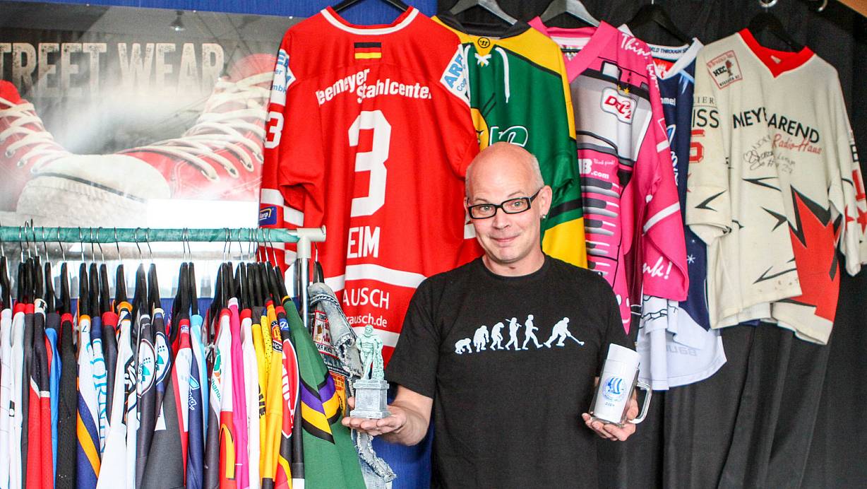 Erinnerungen: Jens Meyer zur Heide steht vor seiner Trikotsammlung. Auf Bügel hängen ihm seine Lieblingstrikots, das HEV-Trikot von Michael Reim sowie die desHC Maustadt, des EHC Bremerhaven, das Weihnachtstrikot der Hamburg Freezers sowie sein erstes Trikot vom Herforder EC. In der linken Hand hält er eine Tasse als Erinnerung an ein Fantreffen. Die kleine Statue in der rechten Hand ist ein Pokal, den Meyer zu Heides Team bei einem Fanturnier gewonnen hat. | © Yvonne Gottschlich