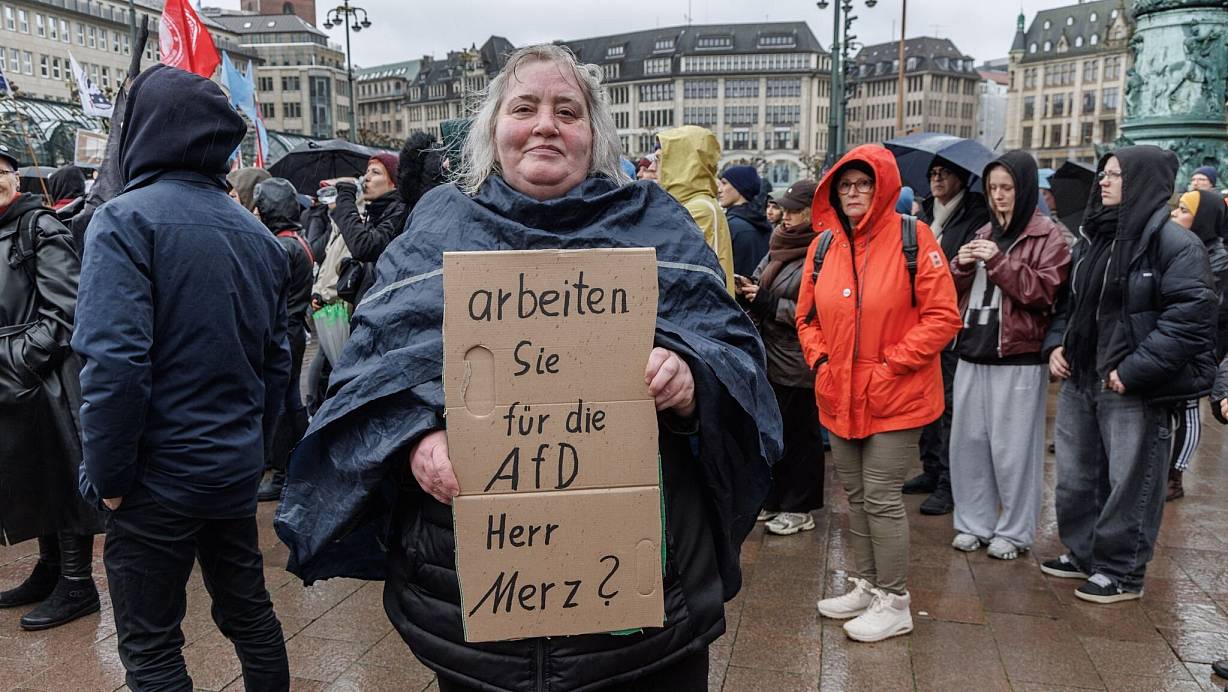 Eine Demonstrantin in Hamburg stellt auf einem Pappschild eine Frage an Bundeskanzler Friedrich Merz (CDU). | © Markus Scholz/dpa