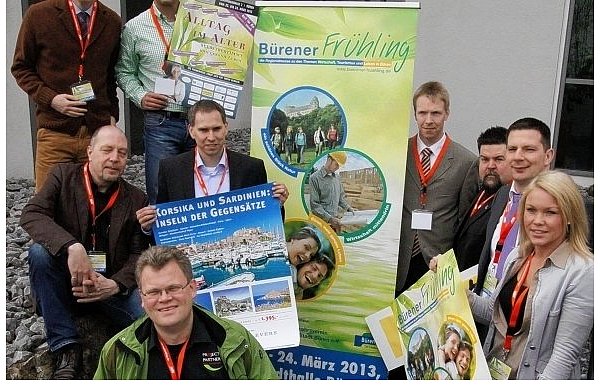 Dirk Kleeschulte (vorn), Ernst Hartung (Gestaltung Messestand Stadt), Jürgen Gausmann, Verkehrsvereinsvorsitzender Ralf Kleine, Wirtschaftsförderer Jan Hüttner, Verkehrsvereins-Vize Marcel Reinecke, Larissa Klegraf (CAT Marketing, Mitte v. l.), Michael Kubat (Stadt Büren) und René Klaas-Jung (hinten v. l.) mit den Ausstellungsplakaten für den Bürener Frühling. - © FOTO: KARL FINKE
