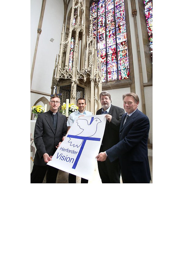(v. l.) Pfarrer Gerald Haringhaus (katholische Gemeinde St. Johann-Baptist), Frank Hölscher, Pfarrer Johannes Beer und Eckhard Wemhöner zeigen vor dem Altar das neue Logo: die Taube auf dem Kreuz sitzend. - © FOTO: KIEL-STEINKAMP