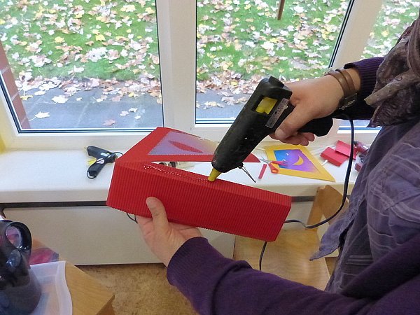 Um die beiden Rautenstücke miteinander zu verbinden, wird nun ein Verbindungsstück aus Wellpapier zugeschnitten: Hierfür empfiehlt es sich, die längste Seite einer Raute doppelt zu nehmen und diese Länge mit einer Breite von 15 Zentimetern aufzumalen und auszuschneiden. Jetzt werden die beiden Längsseiten etwa zwei Zentimeter tief eingeknickt. Danach wird das Verbindungsstück einmal mittig gefaltet. Anschließend werden mit der Klebepistole die geknickten Seiten des Verbindungsstücken an die Innenseiten der beiden Rauten geklebt.