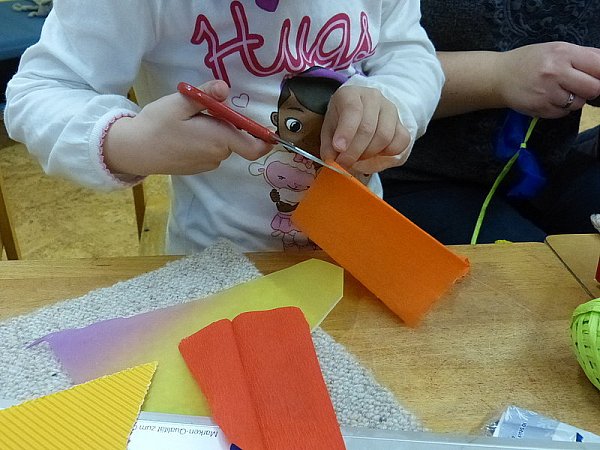 Um den Drachen zu schmücken, werden zum Schluss mehrere kleine Streifen aus buntem Krepppapier ausgeschnitten und jeweils in der Mitte gedreht. Ein Teil der so entstandenen Schleifen werden auf die Spitzen des Drachens verteilt und aufgeklebt. Die restlichen Schleifen werden später zum Schmücken der Schnur benötigt. Mit der Prickelnadel wird zuerst vorsichtig ein Loch in die untere Spitze des Drachens gestochen. Hier wird anschließend die Schnur durchgezogen und von innen verknotet oder verklebt, so dass die Schnur gut hält. Dann werden an der Schnur die übrigen Schleifen befestigt. Das Ende der Schnur wird, damit es nicht beim St. Martinsumzug über den Boden baumelt, an einer Seite festgeklebt. Fertig ist die Laterne.