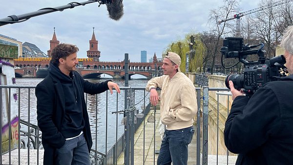 Tommi Schmitt (links) interviewt Robin Gosens in Berlin. Sie stehen am Ufer der Spree, hinter ihnen liegt die Oberbaumbrücke. Neben ihnen steht ein Kameramann, ein Tonarm mit Mikrofon ragt in das Bild. - © ZDF und Gruppe 5