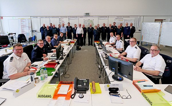 Das Paderborner Team nach Übungsende im Lehrsaalgebäude C des Instituts der Feuerwehr. Im Vordergrund sind die Tische der Sachgebiete 1 bis 4 noch besetzt, während der Rest der Mannschaft vor der großen Lagewand steht. - © Guido Korte/IdF