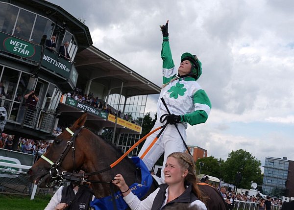 Erinnerung an ihren Vater: Nina Baltromei zeigt nach ihrem historischen Sieg beim deutschen Galopp-Derby mit dem Zeigefinger gen Himmel. - © Marcus Brandt/dpa