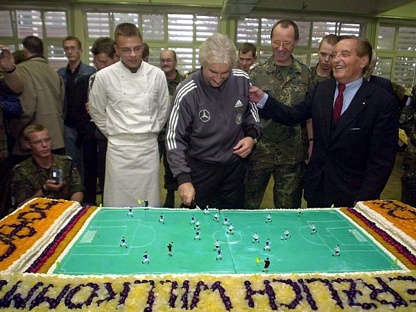 Rudi Völler (M) schneidet beim Truppenbesuch in Sarajevo eine Torte in Form eines Fußallplatzes an. - © Bernd Weißbrod/dpa