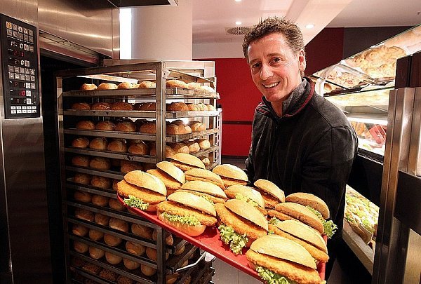 Siegmar Rasche zieht einen Rollwagen mit fertig gebackenen Brötchen aus dem Ofen. Mit Putenschnitzeln und Salat belegt werden sie dem Kunden in der Glasvitrine (rechts im Bild) angeboten. - © FOTO: WOLFGANG RUDOLF