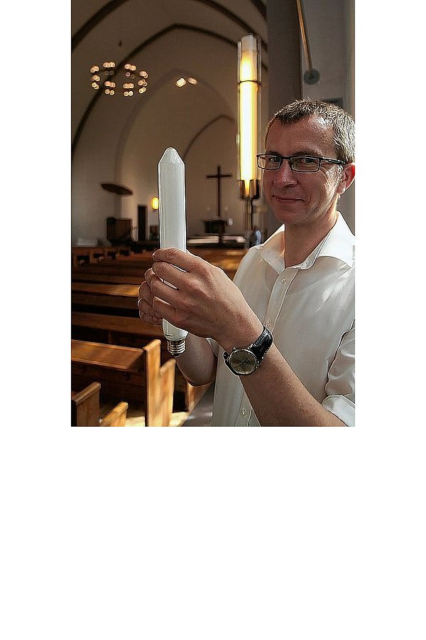 Pfarrer Christian Feuerbaum hält eine der veralteten Leuchtröhren in der Hand. In der Fassung hinter ihm ist der neue Lichtkörper zu erkennen. - © FOTOS: LUDGER OSTERKAMP